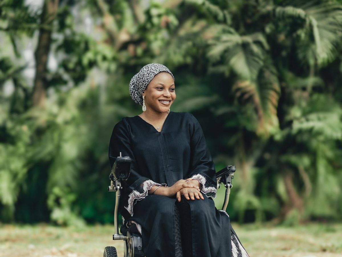 A smiling woman in a wheelchair outdoors among green trees.